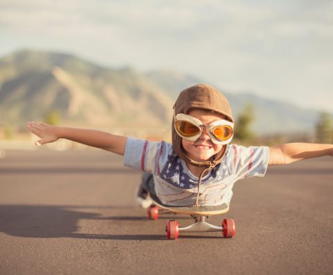 A young boy wearing flying goggles and flight cap outstretches his arms to attempt flying while he rides on his skateboard. He has a large smile across his face as he is imagining taking off.